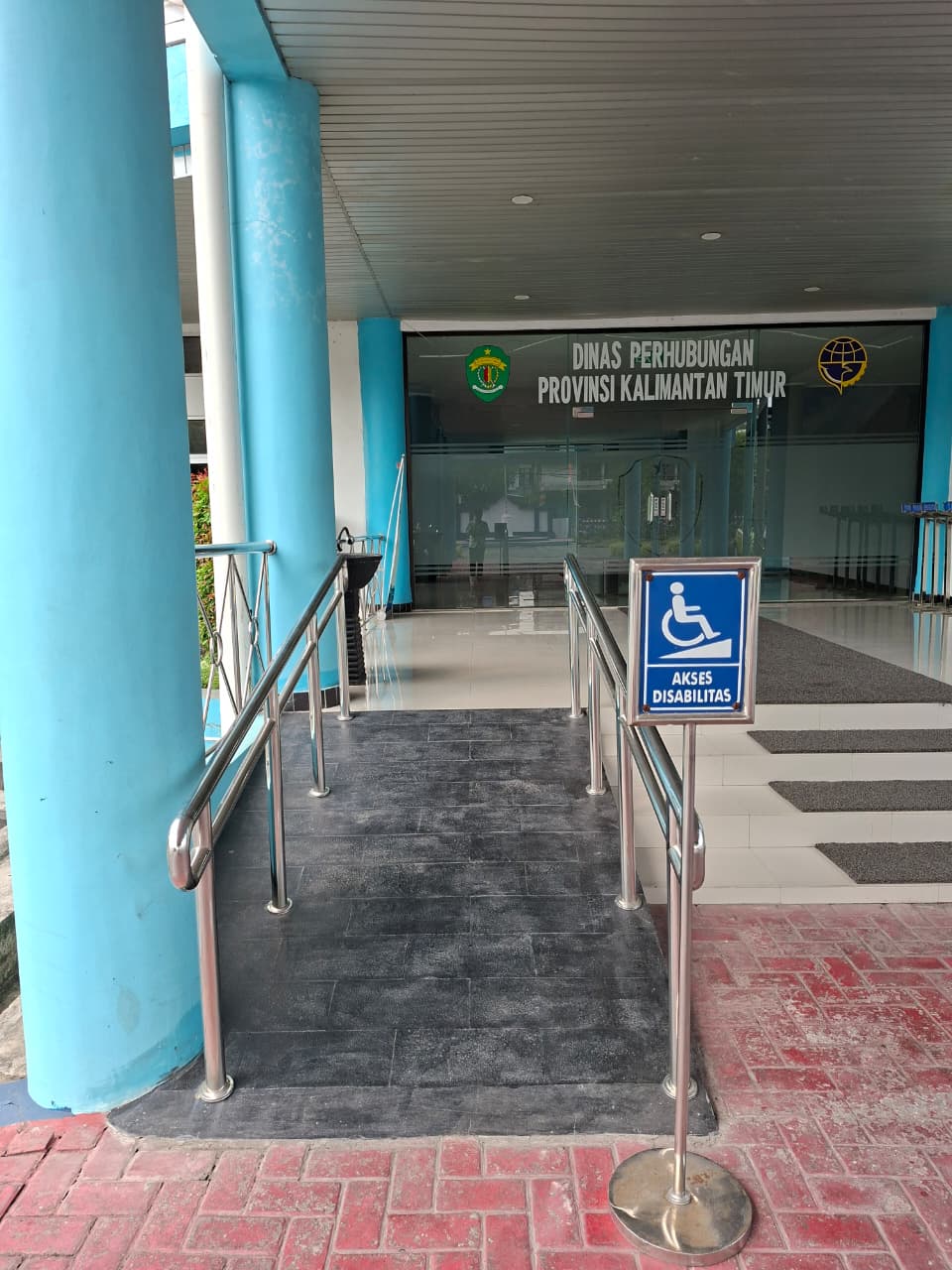 Jalur landai kursi roda berpegangan tangan di kedua sisi, mengarah ke pintu kaca lobi Kantor Dinas Perhubungan Provinsi Kalimantan Timur.